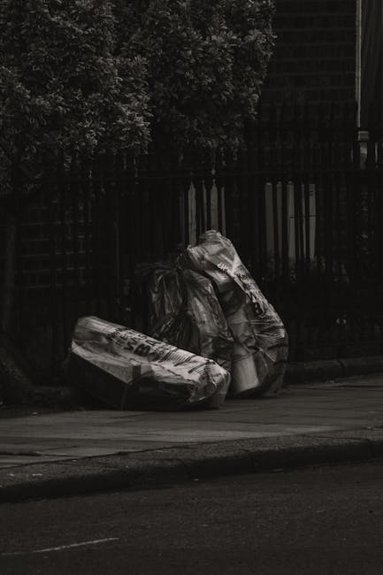 Two large black plastic garbage bags filled with waste are placed on the pavement next to a dark metal fence, which encloses a residential area. One bag appears slightly more crumpled than the other, 