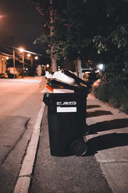 A row of large, wheeled wheelie bins positioned along a pavement beside a brick wall, with the bins arranged in a sequence featuring green, blue, and red colours. The green bin in the foreground is cl