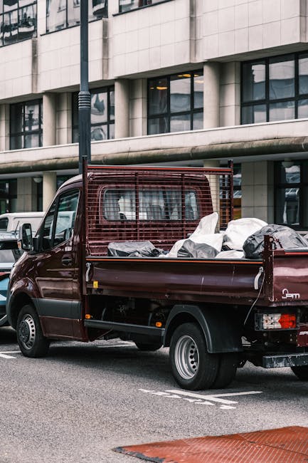 A maroon flatbed truck parked on an urban street in front of a modern concrete building with large glass windows. The truck's bed is loaded with various bags and discarded items, including black and w